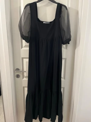 Svart långklänning med puffärmar från Zara - Säljer en svart långklänning från Zara med puffiga, transparenta kortärmar i mesh. Klänningen har en fyrkantig urringning och volang nedtill, vilket ger en cool och trendig look. Perfekt för dig som gillar stilrena och moderna plagg.