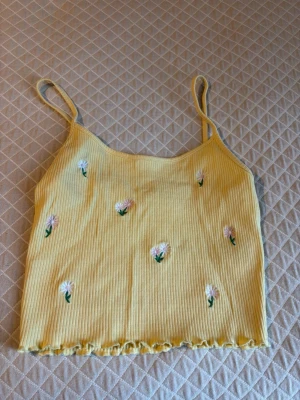 Gul ribbad linnetopp med blommor - Söt ljusgul ribbad linnetopp med tunna axelband och broderade vita blommor med gröna detaljer. Toppen har en croppad passform och vågig kant längst ner. Perfekt för soliga dagar och sommarvibbar.