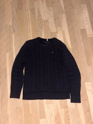 Mörkblå stickad tröja Tommy Hilfiger - Snygg mörkblå stickad tröja från Tommy Hilfiger med klassiskt kabelmönster. Rund hals och långa ärmar, liten logga broderad på bröstet. Perfekt för dig som gillar stilrena och tidlösa plagg med en sportig touch.