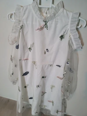 Vit långklänning med blommigt mönster - Superfin vit långklänning med transparenta långärmar och broderade blommor i olika färger, bara axlar. Klänningen har volangdetaljer vid axlar och hals, samt ett skirt lager i mesh. Perfekt för dig som gillar en söt och drömmig stil.