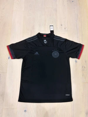 Svart Adidas fotbollströja Tyskland - Säljer en svart Adidas fotbollströja för Tysklands landslag, med diskreta grå ränder på axlarna och tysk flaggdetalj i nacken. Tröjan har Aeroready-material som andas och korta ärmar med röd och gul kant. Perfekt för dig som gillar fotboll och snygg design.