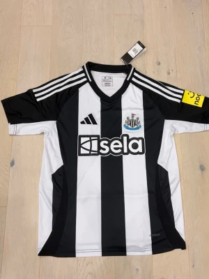 Svarta och vita Newcastle adidas tröja - Säljer en officiell Newcastle United matchtröja från adidas i storlek M. Tröjan har klassiska svarta och vita ränder, klubbmärke på bröstet och sponsortryck. Tillverkad i lätt polyester med AEROREADY-teknologi och slim fit passform. Perfekt för fotbollsfans!
