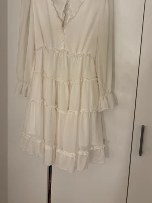 Vit omlottklänning med volanger - Supersöt vit kortklänning med volanger och omlottdetaljer. Klänningen har långärmade transparenta ärmar med volang vid ärmslut, pärlknapp framtill och en öppen rygg med vågiga kanter. Tillverkad i ett lätt, flödande material som ger en romantisk vibe.