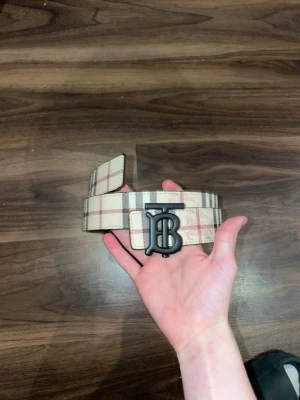 Rutig beige och svart BB bälte med B-spänne. OBS 105cm - Säljer ett brett bälte från Bb med klassiskt rutigt mönster i beige, svart och rött. Bältet har ett stort matt svart B-spänne framtill. Perfekt accessoar för att lyfta vilken outfit som helst.