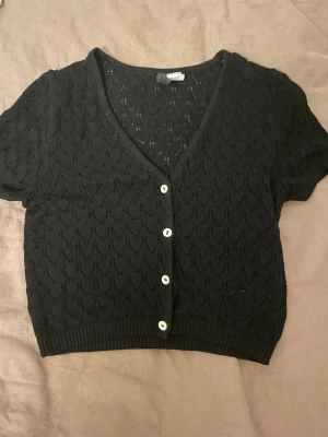 Svart kortärmad kofta från Divided XS - Kortärmad svart kofta från Divided i storlek XS. Tröjan är stickad med ett hålmönster, har v-ringning och vita knappar framtill. Croppad modell med ribbade kanter som ger en snygg och trendig look.