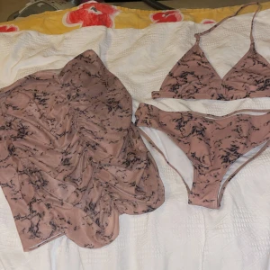 Brun mönstrad 3-delat bikini set - Säljer ett bikini set med svart marmorerat mönster. Kjolen är knälång och har drapering.. Materialet är mjukt och stretchigt, tillverkat av polyester och elastan. Perfekt för strand eller poolhäng i sommar.