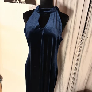 Marinblå långklänning i sammet - Säljer en elegant marinblå långklänning från New Line (Mac Scott) i mjuk sammet. Klänningen har en hög hals med snygg öppning framtill och är ärmlös, vilket ger en stilren och modern look. Perfekt för dig som vill sticka ut med en klassisk och cool vibe.