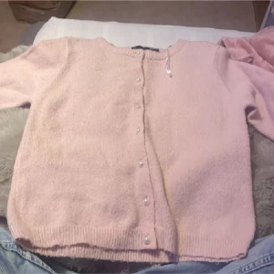 Ljusrosa kofta med pärlknappar 157 Xs - Superfin ljusrosa kofta från 157 i storlek XS. Koftan är stickad och har långa ärmar samt pärlknappar framtill. Perfekt för lager-på-lager och ger en söt touch till din outfit. Rund halsringning och ribbade muddar vid ärmslut och nederkant.