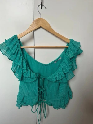 Turkos blus med volanger från Zara - Säljer en turkos blus från Zara med snygga volanger och snörning framtill. Blusen har offshoulder-design och är tillverkad i ett lätt, plisserat syntetmaterial. Perfekt för sommarens alla tillfällen och ger en cool, romantisk vibe.