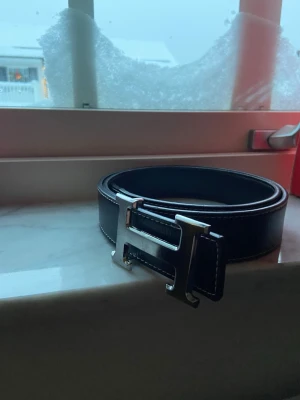 Svart skärp med H-spänne - Snyggt svart skärp med ett stort, silvrigt H-format spänne. Skärpet har vita sömmar längs kanterna som ger en schysst kontrast. Perfekt accessoar för att lyfta din outfit med en stilren detalj.