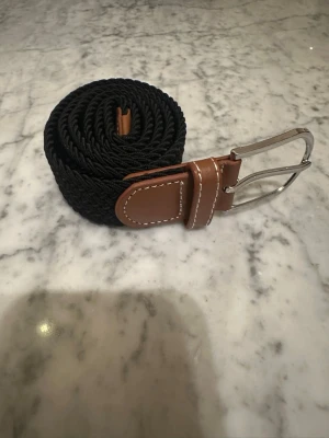 Svart flätad bälte med bruna detaljer - Snyggt svart flätat bälte med bruna skinn-detaljer och vit söm. Spännet är i silverfärgad metall och bältet har en flexibel, vävd struktur som gör det bekvämt att bära. Perfekt för att ge din outfit en stilren touch.