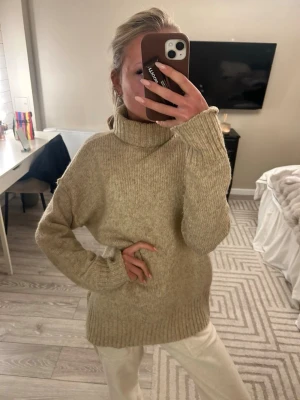 Beige stickad polotröja - Mysig beige stickad polotröja med lång ärm och ribbade muddar. Tröjan har en hög krage och en loose fit som ger en chill och avslappnad vibe. Perfekt att slänga på sig när det är kallt ute.