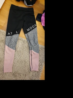 Färgglada leggings från H&M Sport - Snygga leggings från H&M Sport med svart överdel, gråmelerad mitt och ljusrosa ben. De har tryckt text på låren och är perfekta för träning eller chill. Materialet är mjukt och stretchigt syntet för maximal komfort.
