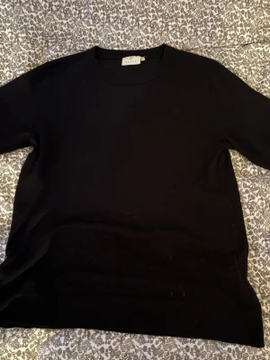 Svart tröja från Kaffe - En stilren svart tröja från Kaffe med rund hals och korta ärmar. Tröjan har en enkel design utan mönster eller detaljer, vilket gör den lätt att matcha med olika outfits. Den har ett stickad material och är inte för tajt eller lös i passformen.