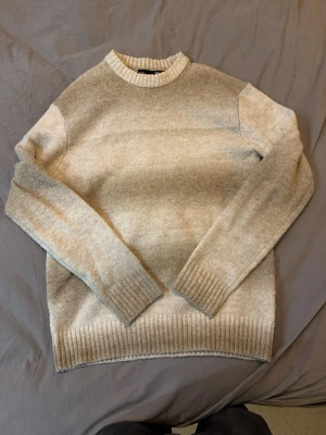 Beige stickad tröja med rund hals - Mysig beige stickad tröja med rund halsringning och ribbade muddar vid ärmslut och nederkant. Perfekt för lager på lager under kyliga dagar. Enkel och stilren design som passar till det mesta.