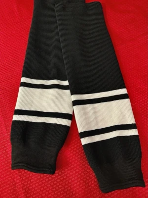 Hockey damasker SR svart M( 43-46) - Säljer ett par svarta ribbade hockey damasker med breda vita ränder och tunna svarta linjer vid bensluten. I mycket bra skick, längd 65cm storlek S-M , 100% polyester 