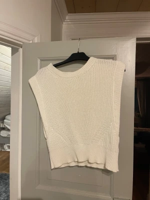 Vit stickad ärmlös tröja från NA-KD - Säljer en vit stickad tröja från NA-KD i storlek S. Tröjan är ärmlös med breda axelpartier och har en rund halsringning. Perfekt att styla över en skjorta eller t-shirt för en trendig look. Ribbad kant nedtill och vid halsen.