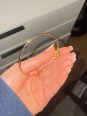 Guldfärgat armband med diamant blomma - Säljer ett tunnt guldfärgat armband med en diamant/sten blomdetalj och en liten sten på ena sidan. Den har en rund form och är enkel att matcha med olika stilar. Perfekt för dig som gillar bling och vill ha något som sticker ut. Tyvärr har en Sten från blomman låssat på den använts mycket!