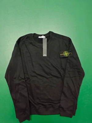 Svart sweatshirt från Stone Island - Säljer en svart sweatshirt från Stone Island med klassisk rund hals och ribbade muddar. Tröjan har den ikoniska Stone Island-loggan på vänster ärm. Perfekt för dig som gillar stilrena och trendiga plagg.