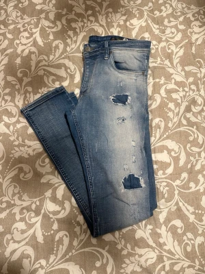 Blå skinny jeans från Jack & Jones - Säljer ett par blå skinny jeans från Jack & Jones med slitna detaljer och hål på framsidan. Modellen är JJORGLEEN ORIGINAL och har klassisk femficksdesign. Jeansen är tillverkade i bomull och har en snygg tvättad look med ljusare partier.