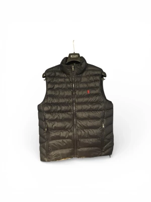 Marinblå dunväst från Polo Ralph Lauren - Marinblå dunväst från Polo Ralph Lauren med horisontella quiltade paneler och röd broderad logga på bröstet. Västen har hög krage, dragkedja framtill och två sidofickor med dragkedja. Perfekt för lager-på-lager och snygg till jeans eller hoodies.