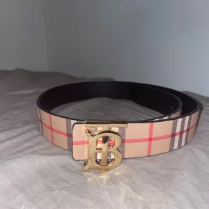 Rutig Burberry bälte med guldfärgat spänne - Säljer ett snyggt Burberry bälte med klassiskt rutig design i beige, rött och brunt. Bältet har ett stort guldfärgat spänne med Burberrys ikoniska B-logga. Insidan är svart och materialet känns lyxigt och robust.
