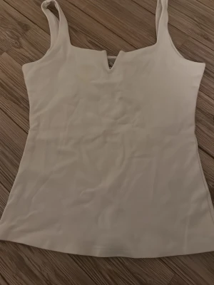Vit linnetopp från H&M med V-detalj - Säljer en stilren vit linnetopp från H&M i storlek S. Toppen har breda axelband och en snygg V-formad detalj vid urringningen. Materialet är mjukt och stretchigt, perfekt för sommardagar eller att styla lager på lager.