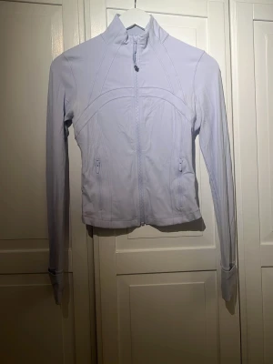Ljusblå träningsjacka från Lululemon - Supersnygg ljusblå träningsjacka från Lululemon med figurnära passform, hög krage och dragkedja framtill. Använd några få gånger och är i bra skick! Tillverkad i stretchigt material med tumhål vid ärmsluten och två fickor med dragkedja. 