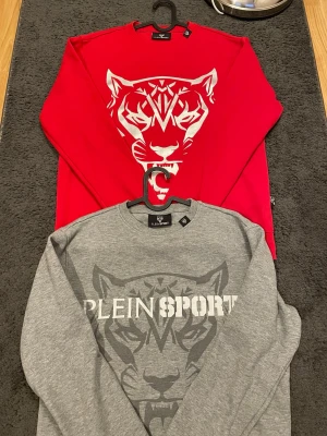 Plein Sport Sweatshirt  - Två snygga sweatshirts från Plein Sport, en i rött med stor vit tiger på bröstet och en i grått med diskret tigertryck och logga. Båda har rund hals, långa ärmar och är gjorda i mjuk bomull. 