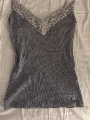 Grå ribbad linnetopp med spets - Säljer en grå ribbad linnetopp från Hollister med smala axelband och v-ringad spetskant i ljusgrått framtill. Toppen är ärmlös och har en slim passform som sitter snyggt mot kroppen. Perfekt för sommarens alla dagar!