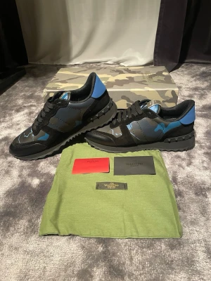 Valentino Rockrunner sneakers blå/svart - Säljer ett par Valentino Rockrunner sneakers i svart och blått med coolt kamouflagemönster. Skorna har snörning, rund tå och platt sula med mönstrad undersida. Materialmix av skinn och textil, med ikoniska nitar på sulan. Perfekt för dig som gillar statement pieces.