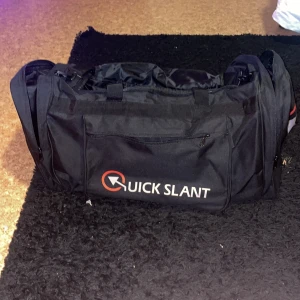 Svart träningsväska Quick Slant - Stor svart träningsväska från Quick Slant med flera fack och dragkedjor. Väskan har bärhandtag och axelrem, samt logga i vitt och rött på sidorna. Tillverkad i slitstark polyester och passar perfekt för gym eller sport.
