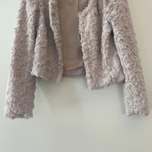 Ljusgrå pälsjacka med croppad passform - Supermjuk och fluffig ljusgrå pälsjacka med croppad modell och lång ärm. Jackan har en cool struktur och är helfodrad för extra komfort. Perfekt för dig som vill sticka ut med en trendig look.