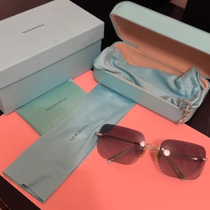 Solglasögon från Tiffany & Co - Snygga solglasögon från Tiffany & Co med stora, ”fyrkantiga” glas och tunna silverfärgade bågar. Sidorna är dekorerade med glittrande stenar för extra lyxig känsla. Kommer med originalask, fodral och putsduk i klassisk Tiffany-blå färg. Lite repor i själva glaset men inget som syns mycket. En Sten är borta, annars otroligt fina. Lite märken på själva stödet. Buda gärna om du är intresserad. 