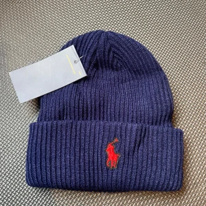 Mörkblå ribbstickad mössa Polo Ralph Lauren - Säljer en mörkblå ribbstickad mössa från Polo Ralph Lauren med klassisk röd broderad logga framtill. Mössan har uppvikt kant och är tillverkad i ett mjukt och värmande material, perfekt för kyliga dagar.