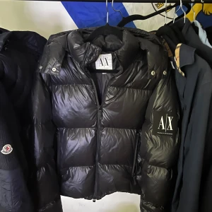 Svart pufferjacka från Armani Exchange - Säljer en svart pufferjacka från Armani Exchange med glansig finish och stor logga på ärmen. Jackan har hög krage, dragkedja framtill och coola detaljer vid axlarna. Perfekt för dig som vill ha en stilren och varm jacka med streetkänsla.