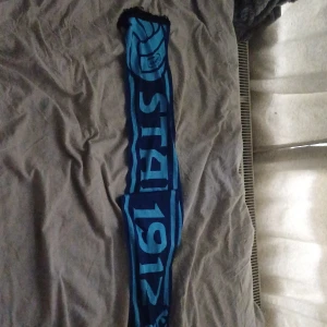 Blå halsduk med text och bollmotiv - Snygg blå halsduk med ljusblå text 'STADION 1912' och ett stort bollmotiv. Halsduken har svarta fransar i ändarna och är perfekt för att visa support på matchen. Materialet känns mjukt och bekvämt mot huden.