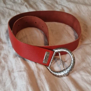 Rött skärp med silverdetaljer  - Säljer ett snyggt rött bälte med en bred rem och ett rundat silverspänne med grafiskt mönster. Bältet har flera hål för justering.ca 95 cm långt från ena sidan till den andra och 84 cm från spännet till mittenhålet.