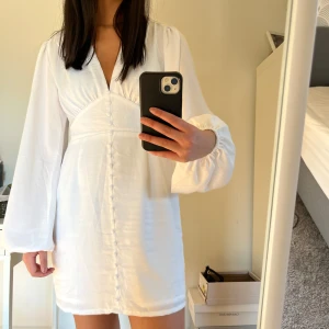 Vit klänning med långa ärmar - Säljer en vit klänning med långa ärmar och v-ringad design. Klänningen har en knappdetalj framtill och en figursydd passform. Prislappen är kvar, för jag fick inget tillfälle att ha på mig klänningen🤍 Nypris 599kr NAKD