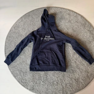 Mörkblå hoodie från Peak Performance - Snygg mörkblå hoodie från Peak Performance med vit logga på bröstet. Tröjan har huva med snörning och en stor magficka framtill. Perfekt för chill eller när du vill ha en sportig look. Långa ärmar och mjukt material.