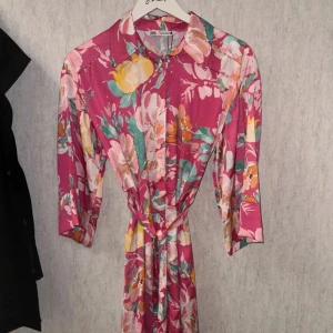 Blommig rosa klänning från Zara - Färgstark blommig klänning från Zara i rosa med inslag av gult, grönt och vitt. Modellen har trekvartsärm, krage, knappar framtill och ett knytband i midjan. Tillverkad i ett glansigt, mjukt material som känns lyxigt och somrigt. Nyskick 