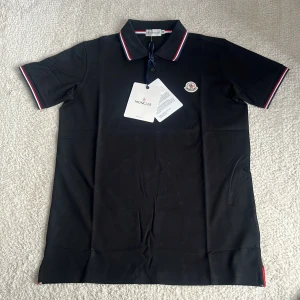 Svart Moncler pikétröja med logga - Svart pikétröja från Moncler med klassisk krage och korta ärmar. Tröjan har Moncler-logga på bröstet och snygga rödvita detaljer på krage och ärmslut. Tillverkad i bomull för en skön och stilren känsla.