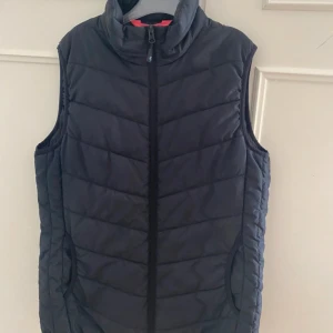 Svart quiltad dunväst med dragkedja - Stilren svart dunväst med quiltad design och hög krage. Västen har hel dragkedja framtill och två fickor på sidorna. Perfekt för lager-på-lager och enkel att matcha med olika outfits. Köpte för 500 kr o aldrig använt 