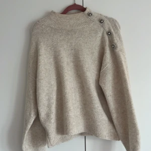 Beige stickad tröja med pärlknappar - Supermysig beige stickad tröja med ribbad hög hals och långa ärmar. Snygga pärlknappar i silver längs ena axeln ger en unik touch. Perfekt för dig som gillar detaljer och vill ha en stilig look.