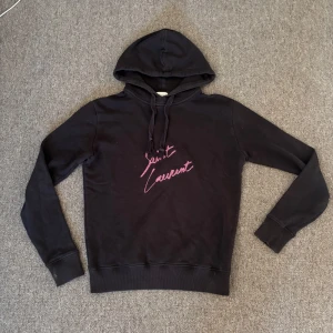 Svart Saint Laurent hoodie med tryck - Svart hoodie från Saint Laurent med huva och dragsnören. På bröstet finns ett rosa broderat Saint Laurent-tryck i cool handskriven stil. Tröjan har långa ärmar och ribbade muddar vid ärmslut och nederkant. Perfekt för en chill och trendig look. Tröjan är sjukt snygg och super bra pris med tanke på att den kostar runt 7000 ny! Storlek xs men kan också passa S