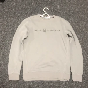 Ljusgrå Sail Racing sweatshirt S - Säljer en stilren ljusgrå sweatshirt från Sail Racing i storlek S. Tröjan har rund hals, ribbade muddar och diskret logga framtill. Perfekt för dig som gillar enkel och clean stil. Materialet är mjukt och skönt, troligtvis bomullsmix.