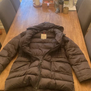 Svart Moncler pufferjacka med huva - Säljer en svart pufferjacka från Moncler med huva och klassisk logga på ärmen. Jackan har dragkedja framtill, två stora fickor och quiltad design. Perfekt för kalla dagar och har en snygg, tidlös look som funkar till allt.