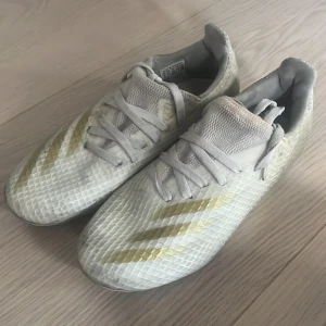 X Ghosted.3 MG Q3 20, fotbollssko - Vit - Säljer ett par Adidas fotbollsskor i vitt med guldiga detaljer och mönstrad ovandel. Skorna har snörning och platt sula med dobbar för bra grepp på planen. Materialet är syntet och designen är sportig med en låg profil. Perfekt för dig som vill sticka ut på fotbollsplanen.