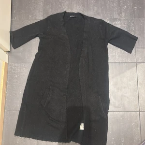 Svart stickad lång kofta  - Svart lång kofta  med trekvartsärm och två fickor framtill. Mjuk och skön känsla, perfekt för dig som gillar enkel och stilren design. Ribbad kant längs öppningen ger en snygg touch.