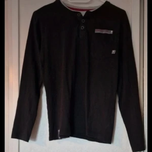 Svart långärmad tröja - Svart långärmad tröja från DNM med rund hals och knappdetaljer framtill. Tröjan är gjord i mjuk bomull och har en enkel, normal passform som funkar till många stilar. Det finns ett litet hål vid lappen bak i nacken.